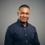 A studio video portrait of an Indian man against a brown background.